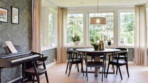 Una habitación con un piano, una mesa de comedor redonda con sillas y ventanas grandes con cortinas.