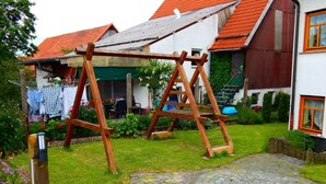 Un ensemble de balançoires en bois dans un jardin, avec un chemin pavé et une maison au toit rouge en arrière-plan.