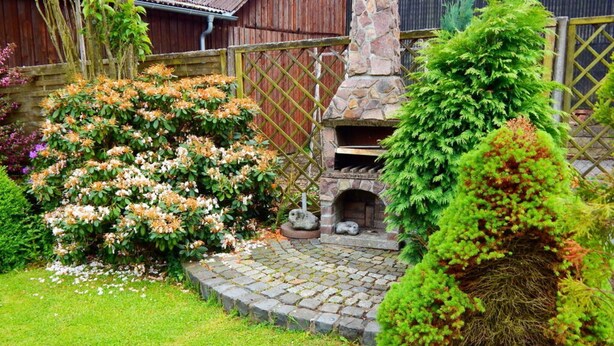 Une cheminée en pierre, entourée d’une végétation luxuriante et de buissons en fleurs, dans un cadre de jardin.