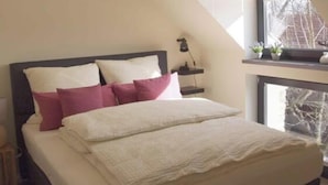 A bedroom with a sloped ceiling, a bed with white and pink bedding, a bedside table with a lamp, and a window with a view of a building. - Holiday Apartment III Bjork (Münchehofe)