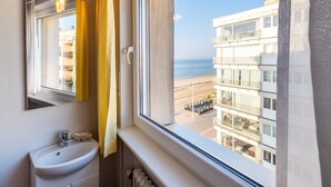 A bathroom with a sink, mirror, and window overlooking a beach and buildings. - Nevada Apartment Near the Beach (Koksijde)