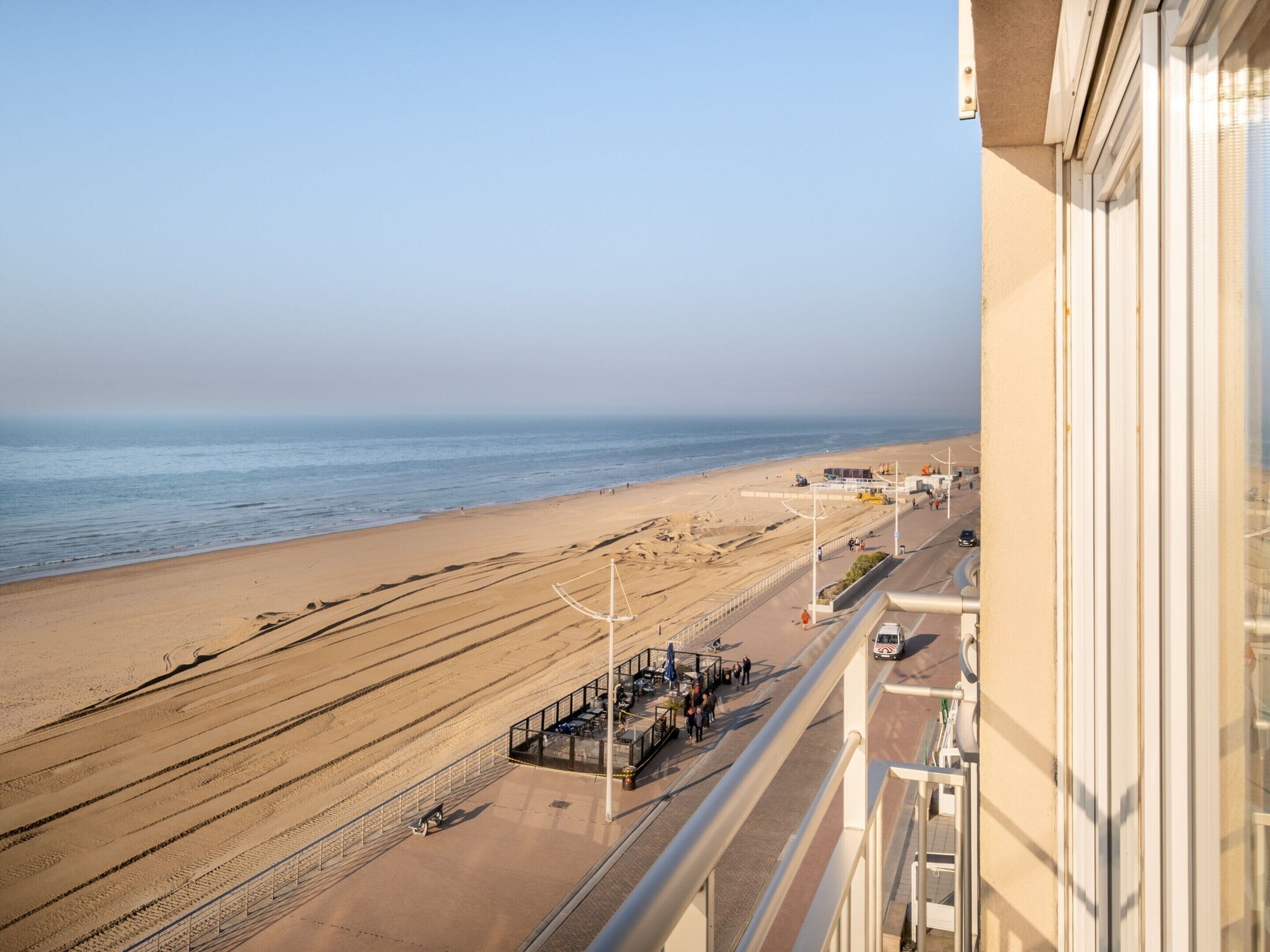A beach view from a balcony overlooking a sandy beach with a promenade, a pier, and a clear sky.