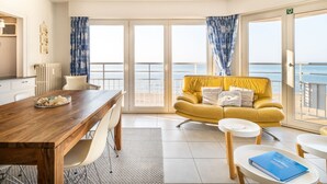 A room with a wooden dining table, a yellow sofa with cushions, a small white table, and a view of the sea through large glass doors. - Nevada Apartment Near the Beach (Koksijde)