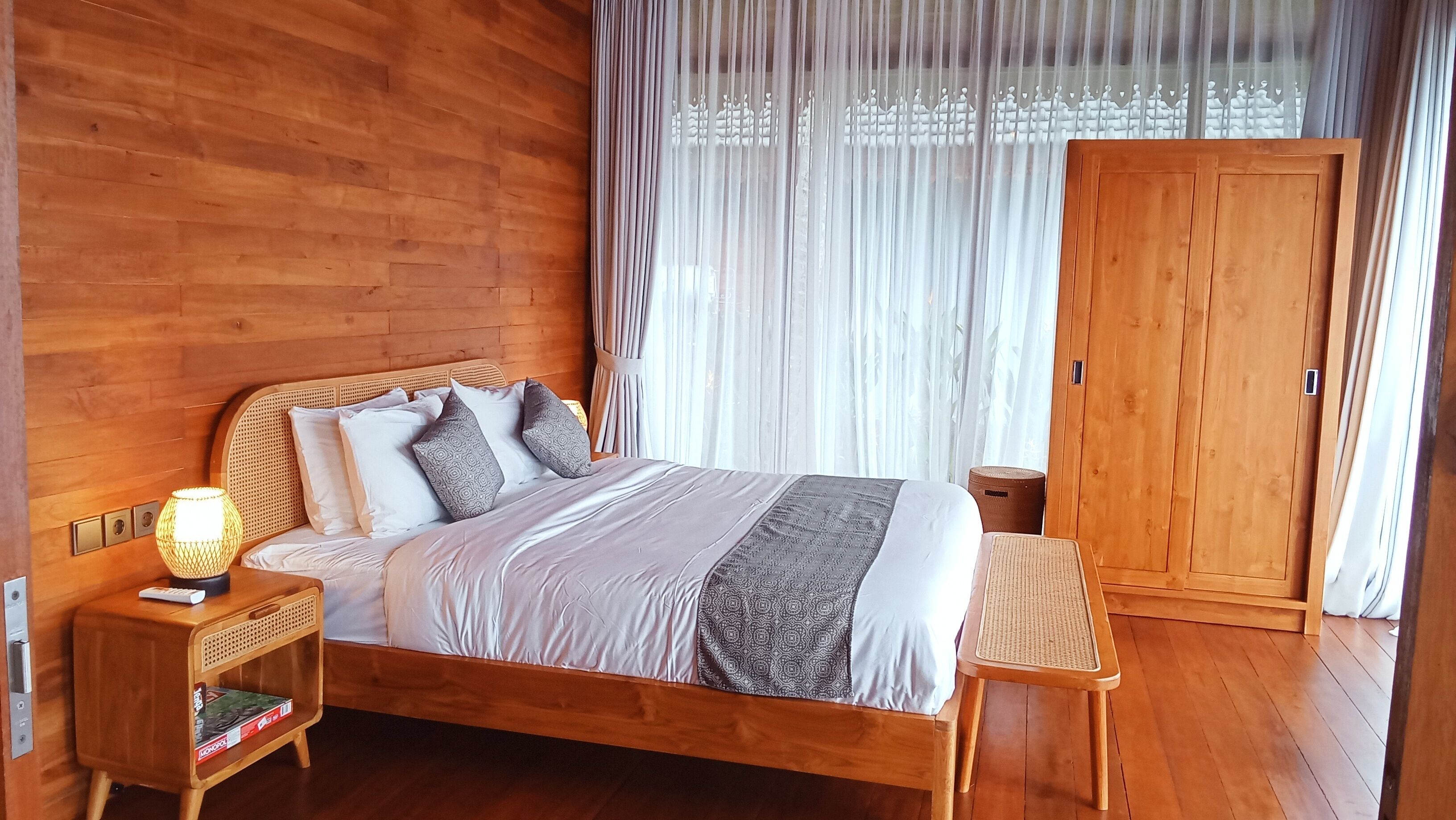 A wooden room with a bed, a nightstand, a lamp, a wardrobe, and a window with sheer curtains.