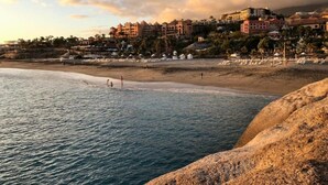 A coastal beach scene with a rocky foreground, a sandy beach, and a row of buildings in the background.