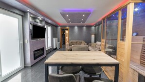 A modern living room with a wooden dining table, grey sofas, and a TV mounted on the wall. - Holiday Home in Verviers With Sauna (Verviers)