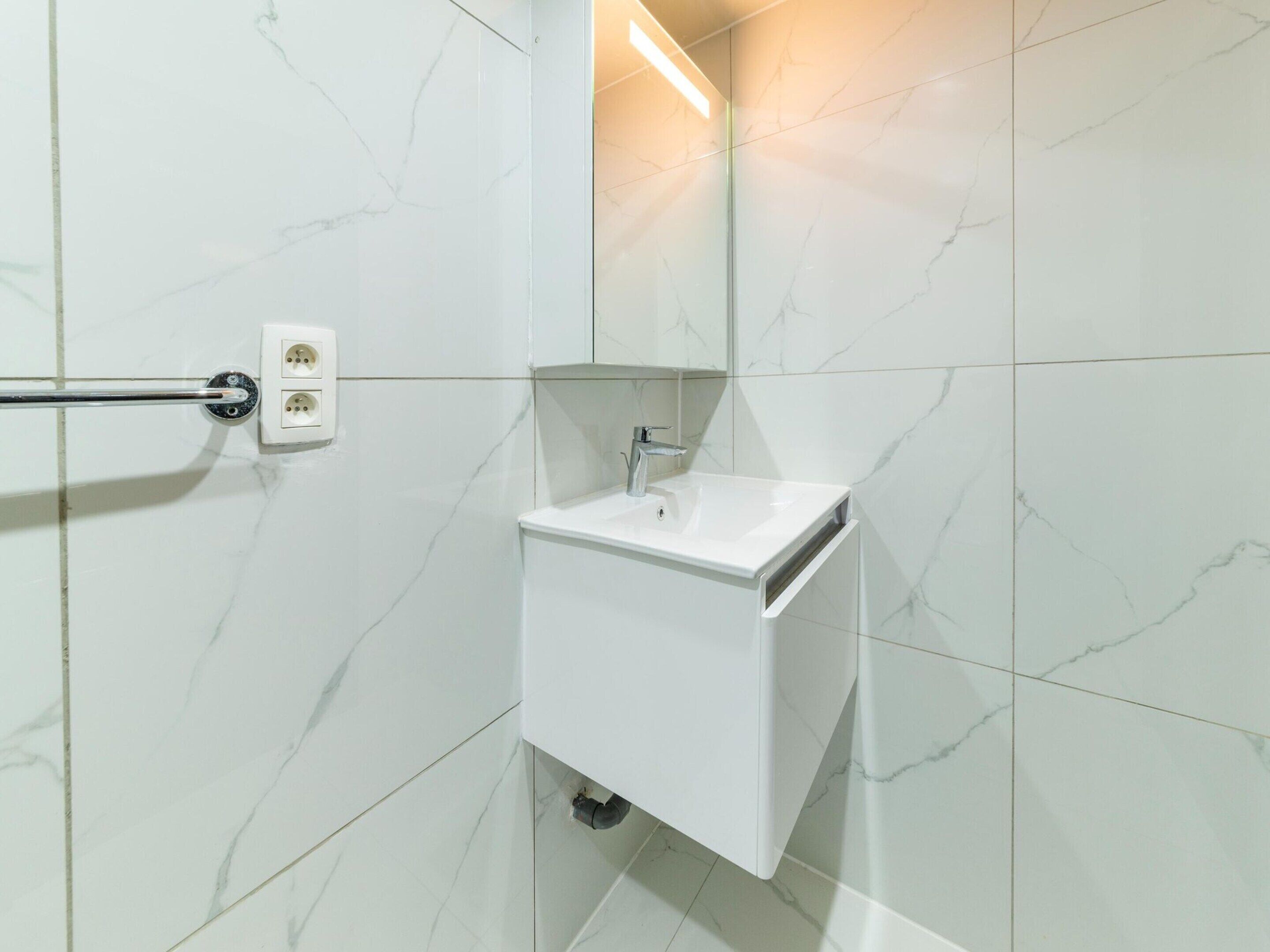 Ein modernes Badezimmer mit weißen Marmorfliesen, einem weißen Waschbecken und einem Spiegel darüber.