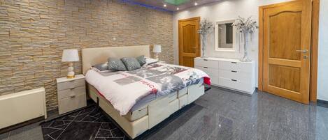 Quarto moderno com parede de pedra, cama com colcha estampada em padrão geométrico, mesa de cabeceira com abajur, cômoda e porta de madeira.