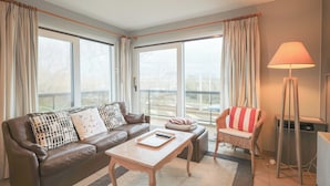 A living room with a brown leather sofa, a wooden coffee table, a wicker armchair, and a floor lamp. - Apartment in Belgium Near Coastal Charm (Koksijde)
