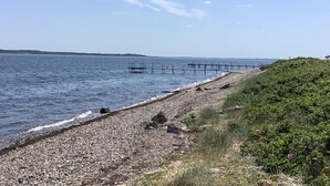 A pebbly beach with a wooden pier extending into the water, a grassy hill on the right, and a clear sky.