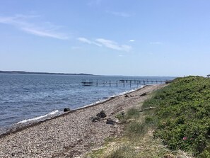 A pebbly beach with a wooden pier extending into the water, a grassy hill on the right, and a clear sky.