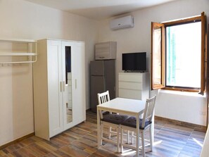 Ein kleiner, gut beleuchteter Raum mit einem weißen Tisch, Stühlen, einem Fernseher, einem Kühlschrank und einem Fenster mit Blick auf Gebäude.