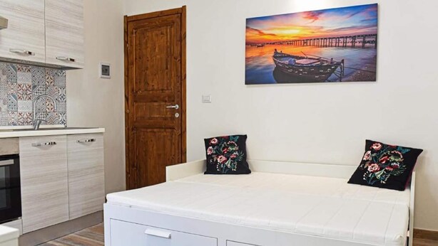 A bedroom with a bed, a wooden door, a kitchen area, and a painting of a sunset over a pier. - Pleasant Apartment in Alghero (Alghero)