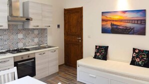 A compact kitchen with a stove, sink, and white cabinets. There's a wooden door and a painting of a sunset over a pier. - Pleasant Apartment in Alghero (Alghero)