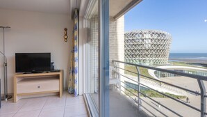 Un balcón con televisor, cortinas y vistas a un edificio moderno junto al mar.