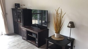 A living room with a dark wooden entertainment center, a flat-screen TV, a small black table with a lamp, and a decorative plant. - Holiday Apartment Moby Dick (St. Peter-Ording)