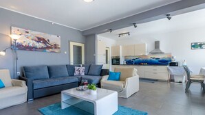 Ein modernes Wohnzimmer mit einem grauen Sofa, einem weißen Couchtisch und einem blauen Teppich. Im Hintergrund ist eine Küche mit weißen Schränken und einer blauen Arbeitsplatte zu sehen.