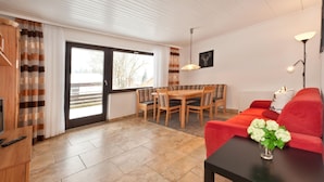 En moderne stue med en rød sofa, et spisebord med stole, et fjernsyn og et stort vindue med gardiner.