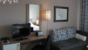 Een hotelkamer met een televisie, bureau, stoel, bank, spiegel, wandlamp en een ingelijst schilderij.