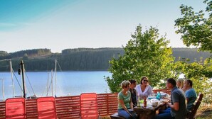 Een groep mensen zit aan een houten tafel buiten, met een meer en bos op de achtergrond.
