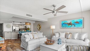 A spacious living room with a white sofa set, a coffee table, and a patterned ottoman. There is a ceiling fan, a painting of sea turtles, and a kitchen area in the background. - BREATHTAKING VIEW! 3BR/2BA Condo Unit 8B Centrally Located In North Myrtle Beach (North Myrtle Beach)