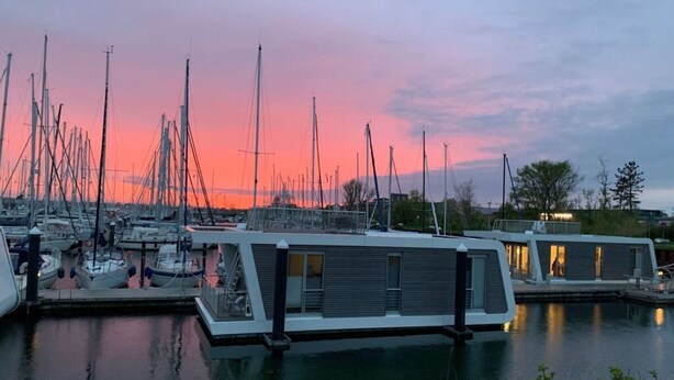 Een jachthaven met meerdere zeilboten afgemeerd, een modern gebouw met grote ramen, en een rustig wateroppervlak dat de lucht weerspiegelt.