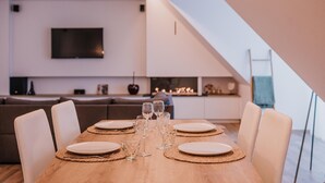 Ein zeitgemäßer Essbereich mit einem Holztisch, gedeckt für vier Personen, umgeben von weißen Stühlen. Im Hintergrund ist ein Fernseher an der Wand befestigt, und rechts ist eine Treppe zu sehen.