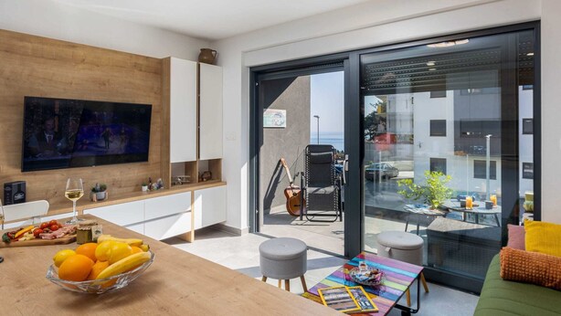 A modern living room with a large flat-screen TV, a wooden dining table with fruit and wine, a colorful coffee table, and a sofa with cushions. - Apartment Good Vibes (Konavle)