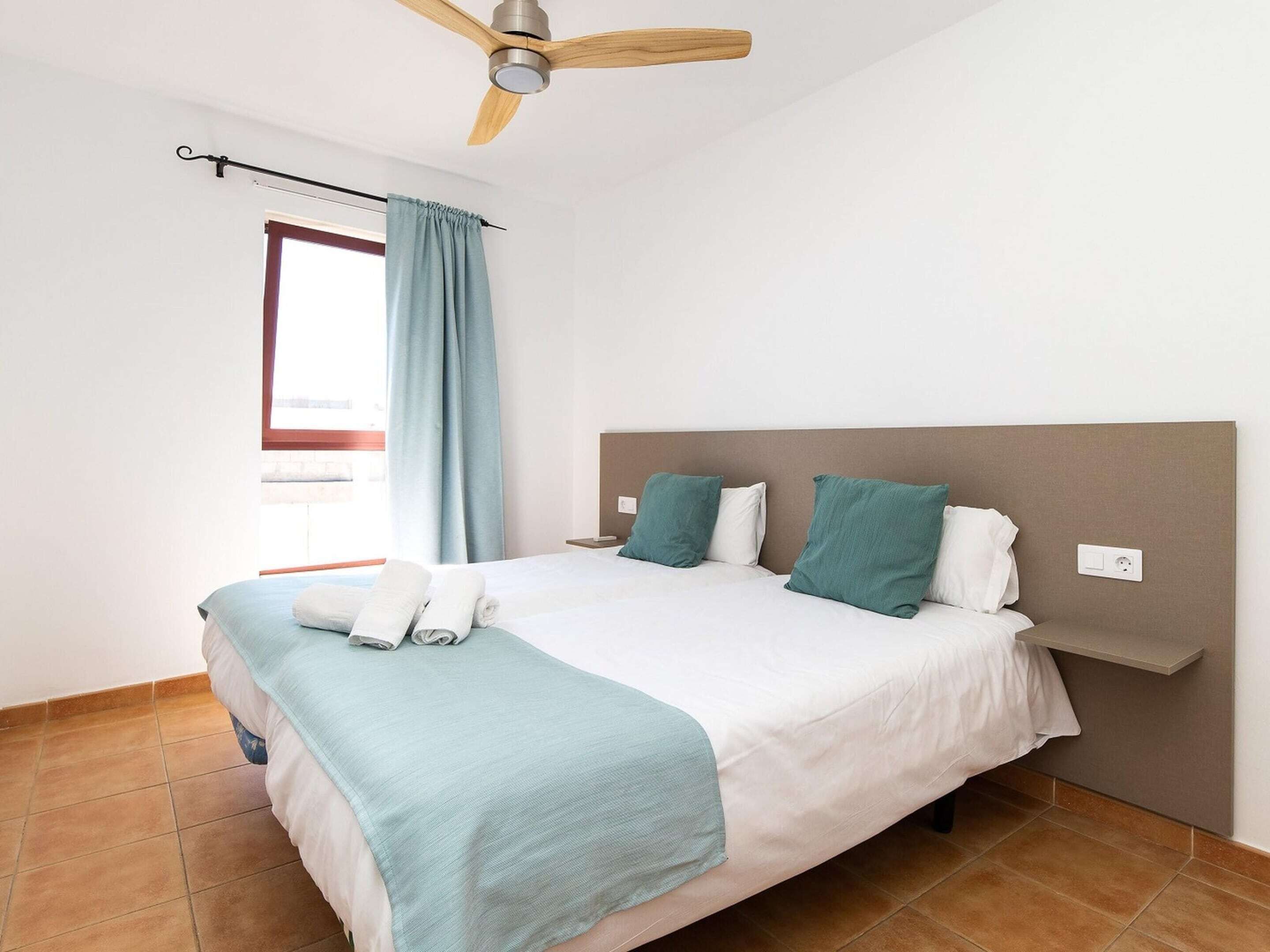 A bedroom with a bed, a ceiling fan, a window with curtains, and a wall-mounted shelf.