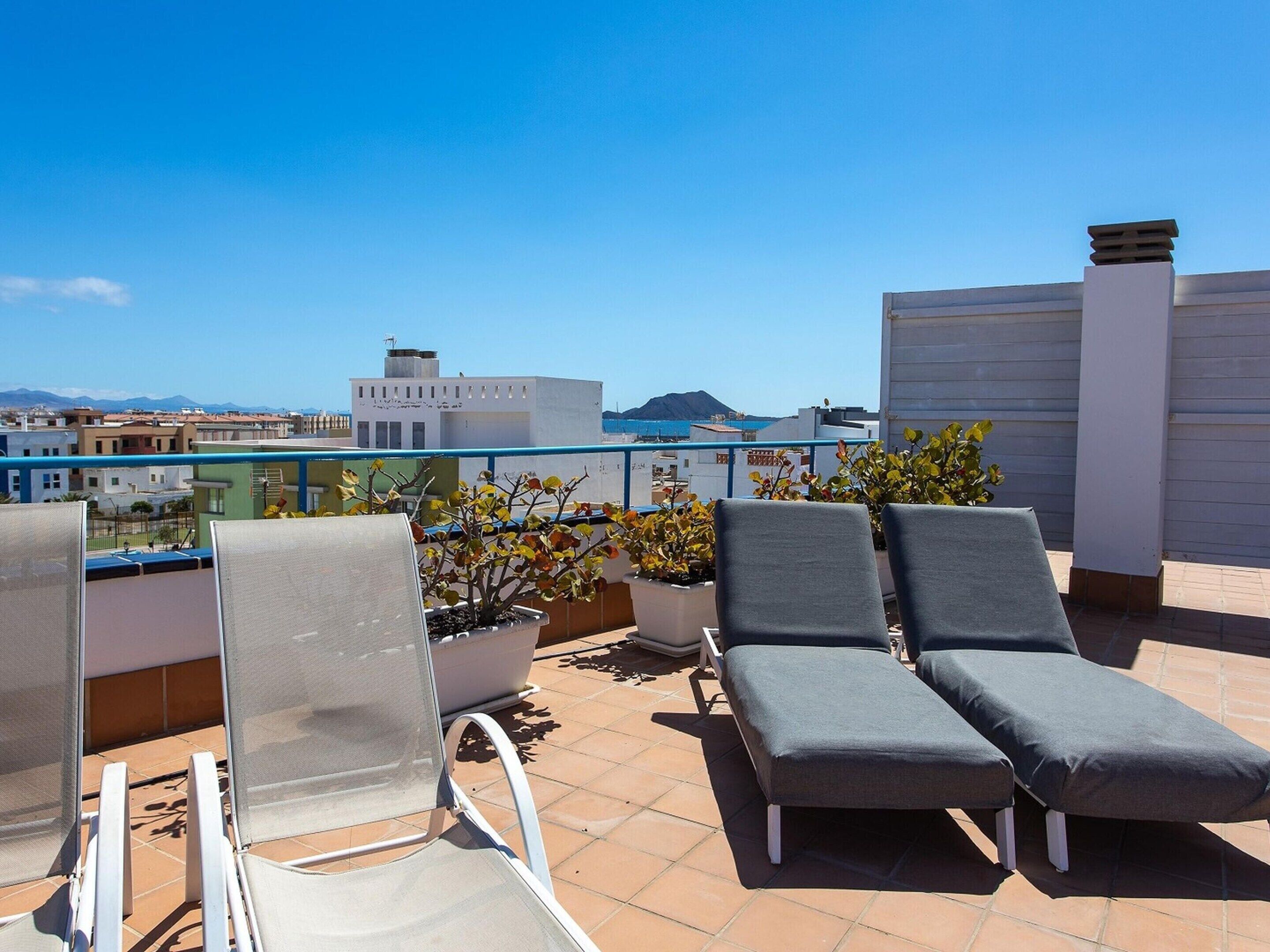 A rooftop terrace with two lounge chairs, a potted plant, and a view of the city and sea.