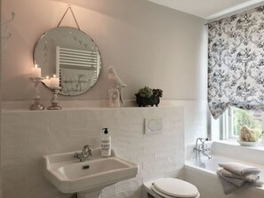 Une salle de bain équipée d’un lavabo, de toilettes et d’une baignoire. On y trouve un miroir au-dessus du lavabo, un distributeur de savon fixé au mur et une fenêtre ornée de rideaux à motifs floraux.