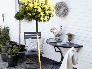Un espace jardin aménagé avec un banc, des plantes en pot, une statue de coq décorative et une table avec des tasses et des soucoupes.