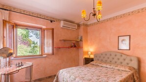 Un dormitorio con cama, mesita de noche, espejo, ventana con cortinas, estante con libros y aire acondicionado instalado en la pared.