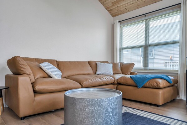 A tan leather sectional sofa with pillows, a round metal table, and a window with blinds. - Waterfront, King Suite, Fully Stocked, Minutes from New Orleans, Pets Welcome (Barataria)