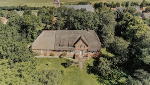 Eine Vogelperspektive eines großen, historischen Hauses mit einem markanten Turm, umgeben von üppiger Vegetation und Feldern.