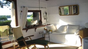 Ett mysigt rum med en gungstol i trä, en soffa, ett litet bord med en vas och ett fönster med utsikt över grönskan.