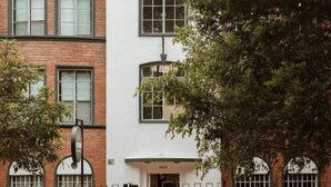 Un bâtiment à deux étages, doté d’une façade blanche et d’un rez-de-chaussée en briques. Chaque étage compte plusieurs fenêtres, dont certaines sont équipées de volets. Un arbre se trouve devant le bâtiment, et un taxi jaune passe dans la rue.
