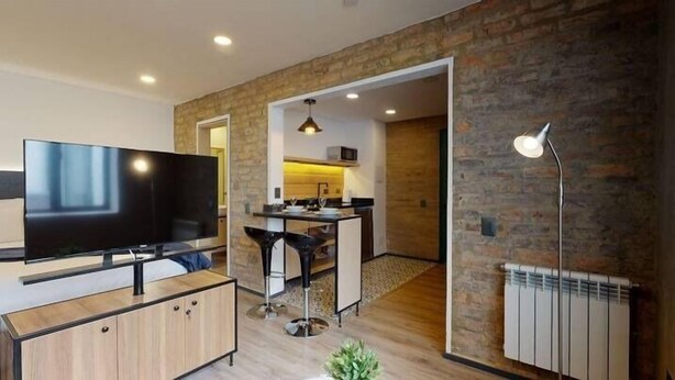 Un salon moderne doté d’un téléviseur à écran plat, d’un meuble en bois, d’un coin repas avec un tabouret de bar et d’une cuisine équipée d’un four à micro-ondes.