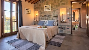 Une chambre avec un mur en pierre, un lit avec une tête de lit, une table de chevet, une commode, une fenêtre avec des rideaux et une porte donnant sur une autre pièce.