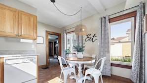 Una cocina con armarios de madera, un lavavajillas blanco, una mesa de comedor con cuatro sillas blancas y una lámpara colgante.