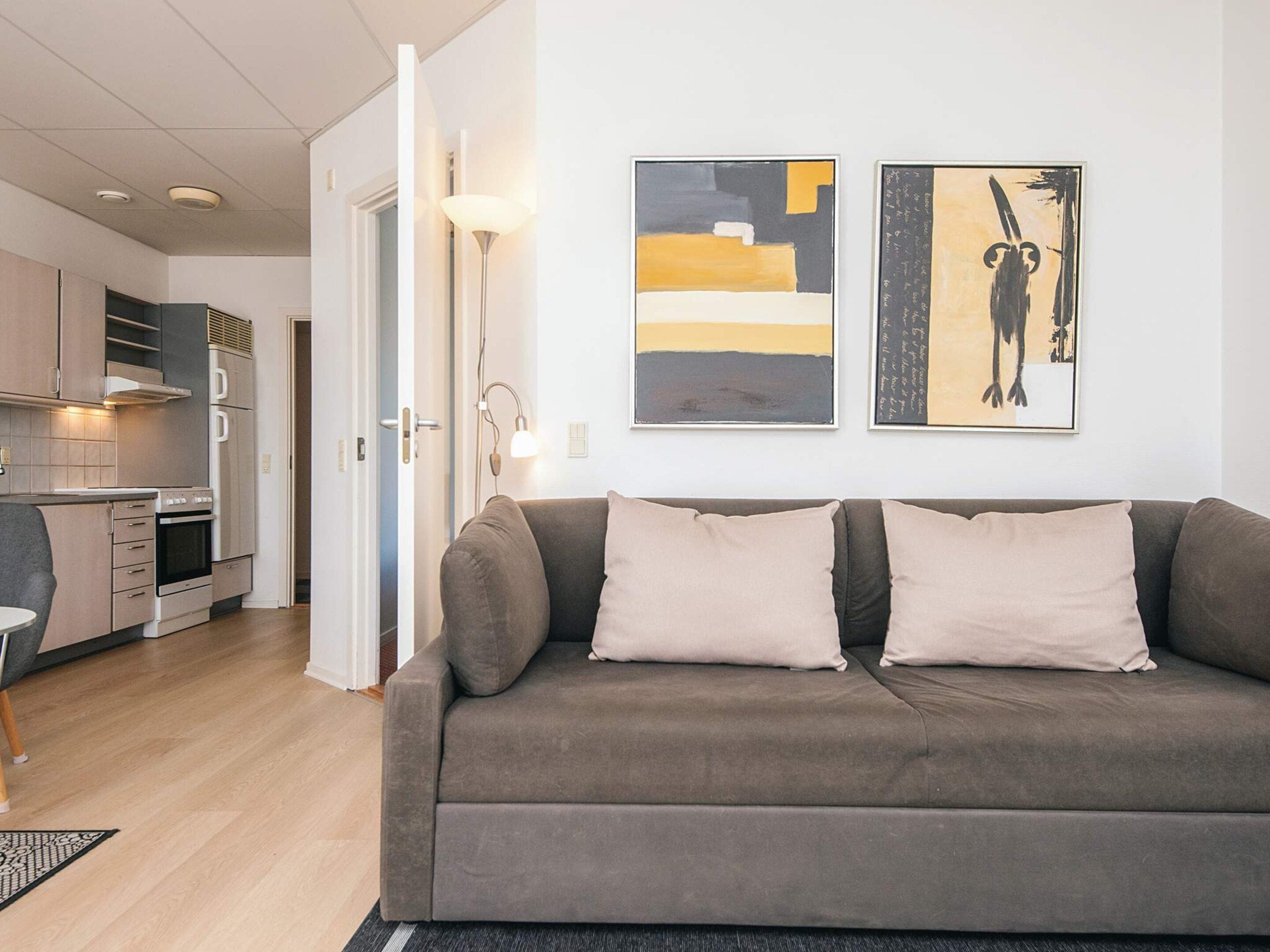 A modern living room with a grey sofa, a kitchen in the background, and two abstract paintings on the wall.