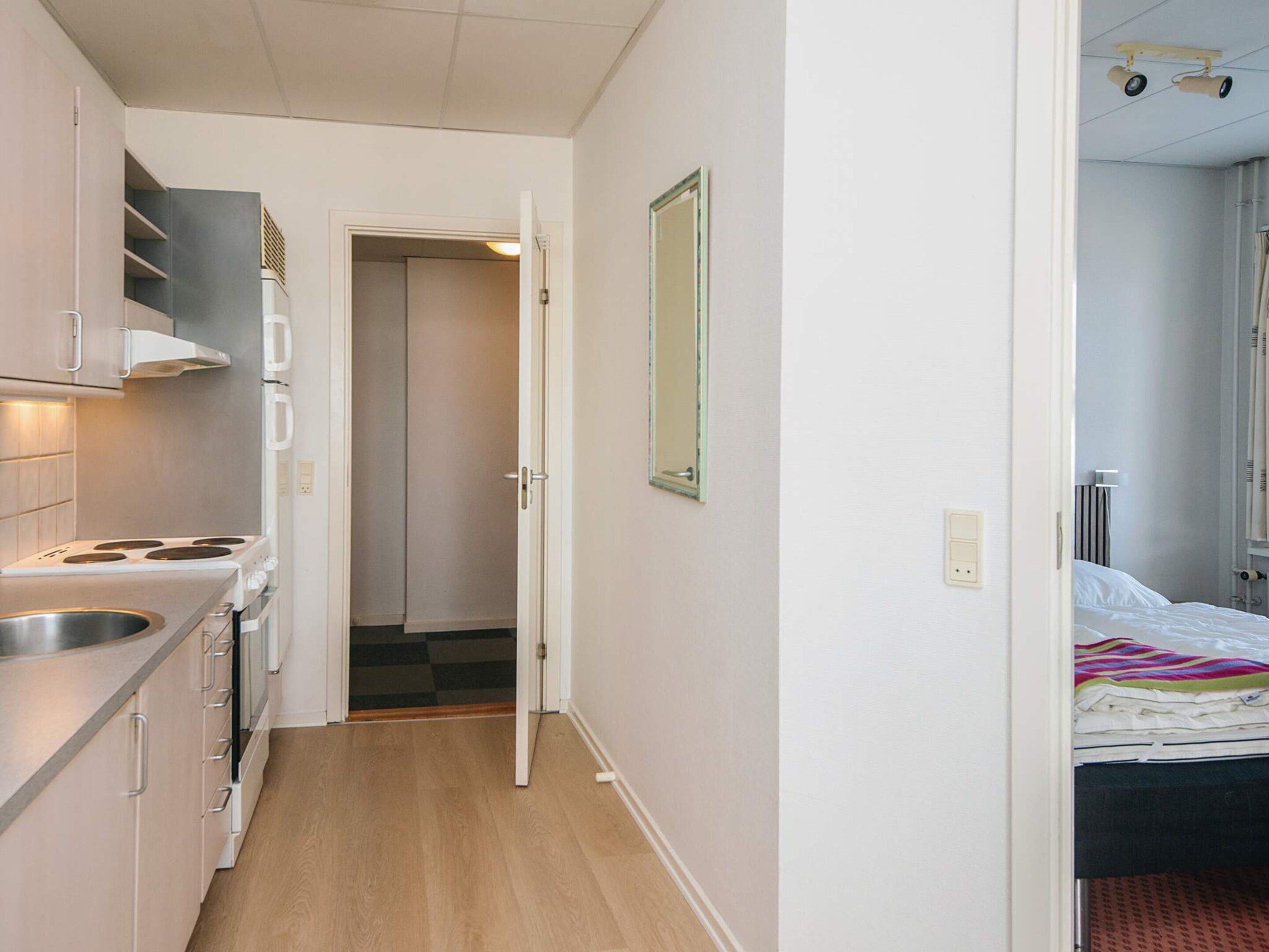 A compact kitchen with a stove, sink, and cabinets, a hallway leading to a bedroom with a bed and a window.