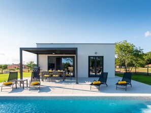 Un área de piscina moderna al aire libre con un porche cubierto, sillas de descanso y una parrilla.
