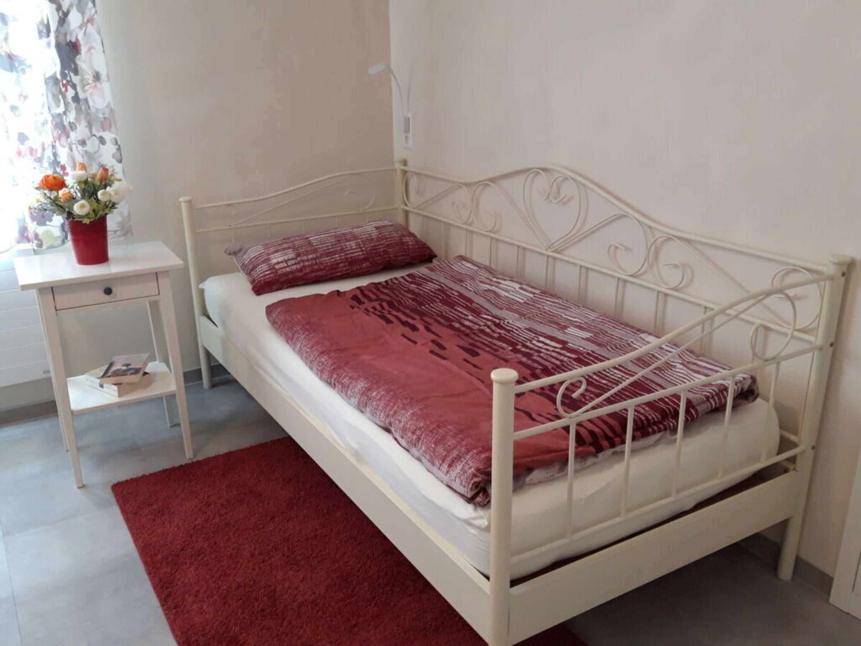 A single-bed room with a white metal frame, a red patterned bedspread, a red rug, a small bedside table with a red vase, and a window with floral curtains.