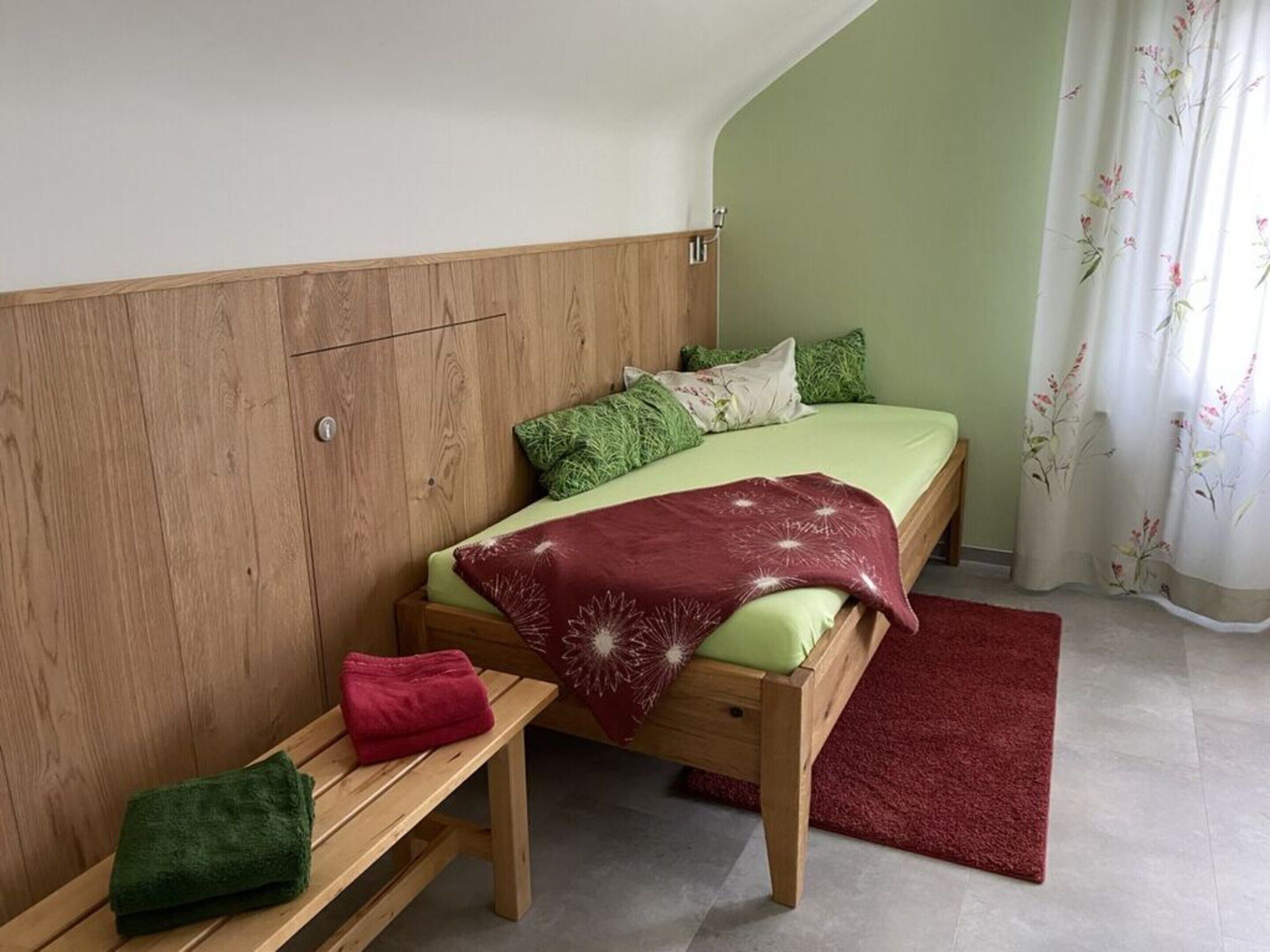 A small, cozy bedroom with a wooden headboard, a single bed with green bedding, a red throw blanket, and a bench with a green towel.