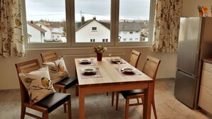 Ein Esszimmer mit einem Holztisch, gedeckt für vier Personen, mit Stühlen mit floralen Kissen. Eine Kühlstelle befindet sich rechts, und das Zimmer verfügt über ein großes Fenster mit Vorhängen.