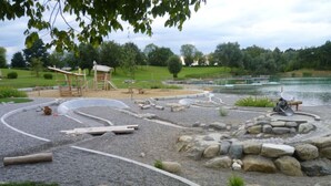 Ein Park mit einem Spielplatz, einem Teich und einer Sitzgruppe aus Stein.