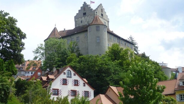 Ein schlösserartiges Gebäude mit einem markanten Turm, umgeben von üppiger Vegetation und Wohnhäusern.