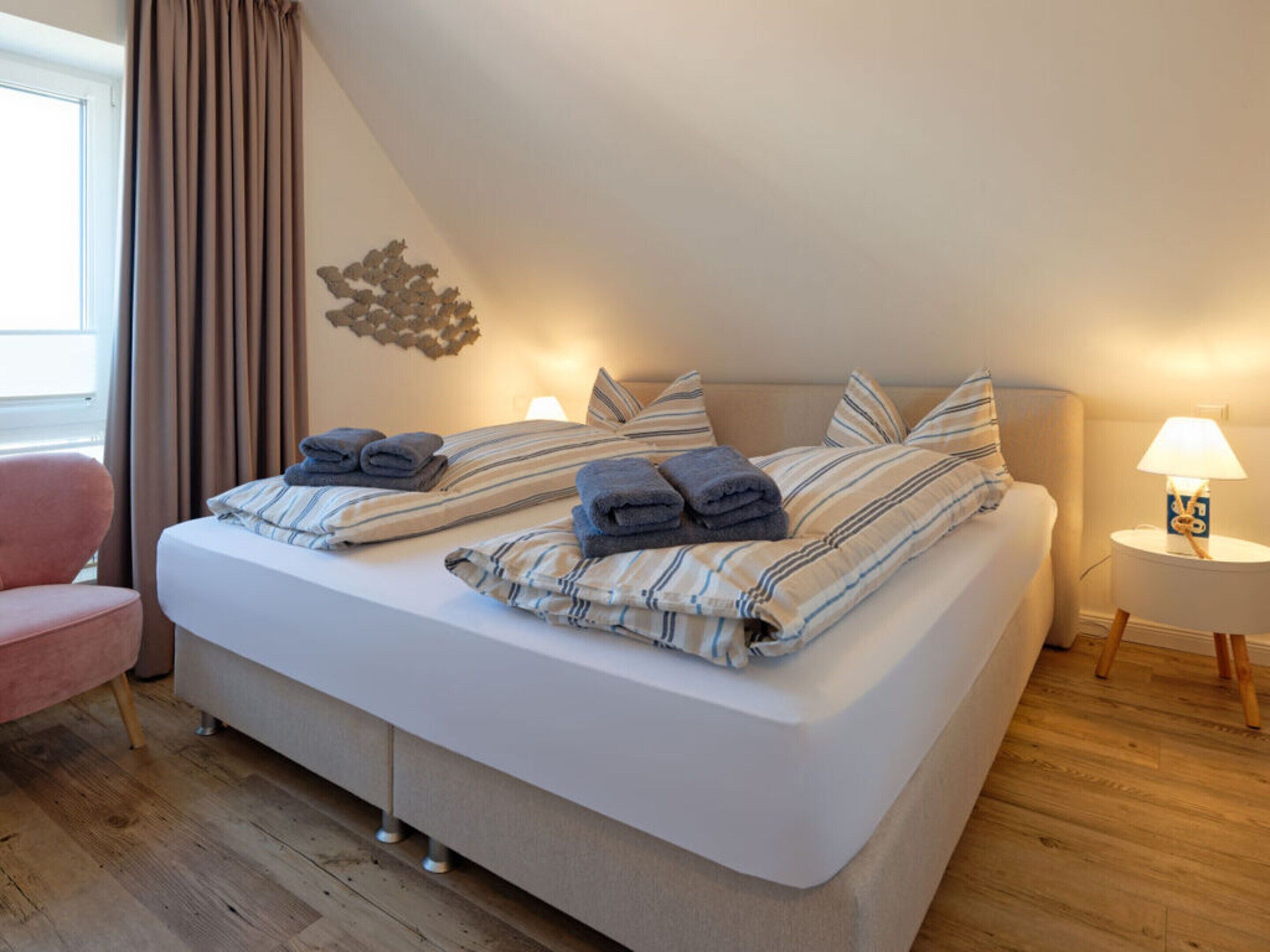 A neatly made bed with striped bedding and folded towels, a bedside lamp, and a pink chair in a room with wooden flooring and a window with curtains.