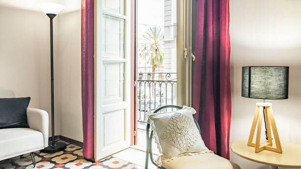 Una habitación con un sofá blanco, una silla con un cojín de punto, una mesita redonda con una lámpara y vistas a un balcón con una palmera.